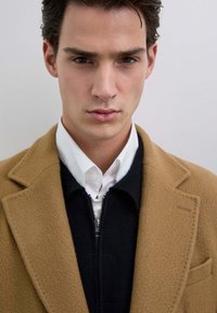 Joven con cabello oscuro que lleva una camisa blanca con cuello, un suéter azul marino con cremallera y un abrigo de lana color camello, mirando hacia adelante.