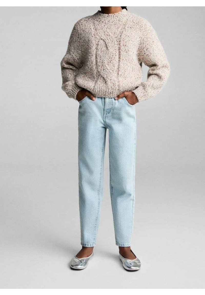 Personne portant un pull beige à torsades, un jean taille haute bleu clair et des ballerines argentées, les mains dans les poches, debout sur un sol gris.