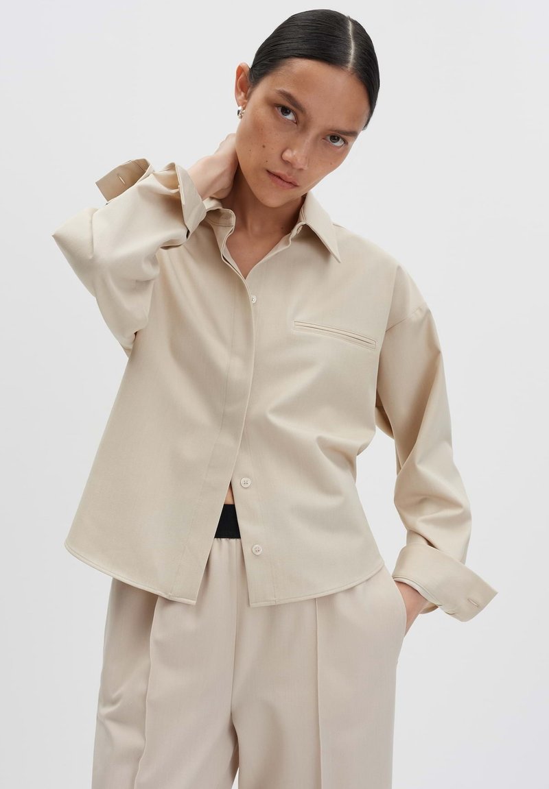 Donna con i capelli scuri in una camicia oversize beige con bottoni e pantaloni larghi abbinati, una mano in tasca, su uno sfondo leggero e semplice.