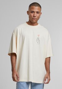 Kremfarget oversized t-skjorte med rund halslinning. Har et enkelt grafisk motiv av en hånd som lager et hjertegrep på brystet.