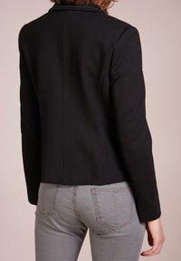 Černá zkrácená bunda s hladkou texturou, zdobená decentním límcem, štíhlým siluetou a precizním štepováním. Pod ní jsou viditelné šedé džíny.