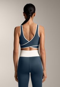 Temno modra športna modrc za aktivne aktivnosti z belimi detajli, ki se ponaša z obliko križanja na hrbtu in širokim belim pasom. Gladek, raztegljiv material.