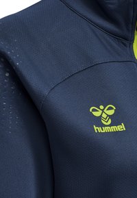 Chaqueta deportiva azul marino con tejido texturizado, con un logo "hummel" amarillo y un patrón de puntos a lo largo del hombro para ventilación.