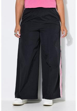 Person iført sorte bukser med vide ben med pink og hvide sidelister, en pink top, hvide sneakers og ringe på begge hænder.