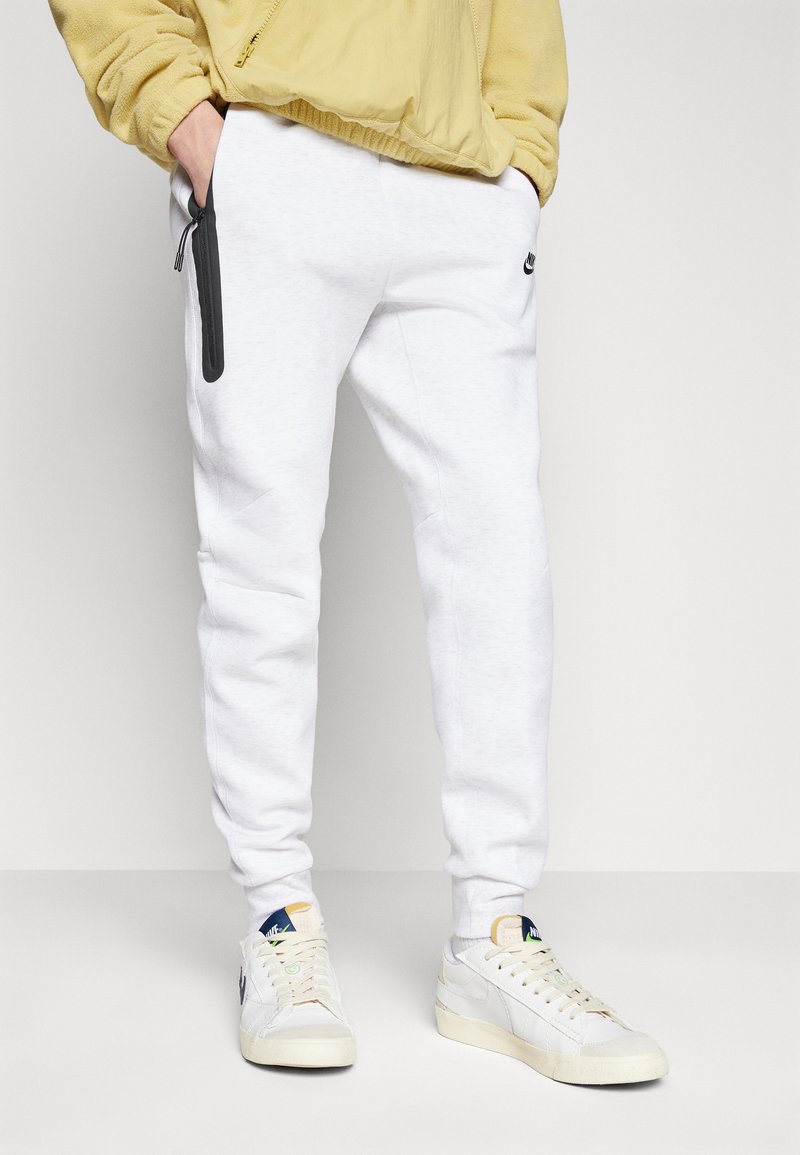 Pantalones de chándal blancos con bolsillos con cremallera negra, corte slim, combinados con zapatillas beige y blancas. La parte superior es una chaqueta ligera amarilla, con cremallera.