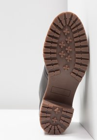 Semelle d'une botte marron avec des crampons épais et à motifs présentant des rainures en forme d'étoile et rectangulaires reposant contre un coin blanc.