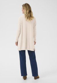 Cárdigan largo beige con tejido texturizado, un corte holgado y lados con aberturas, combinado con jeans de pierna ancha azul oscuro y botas de tobillo marrones.