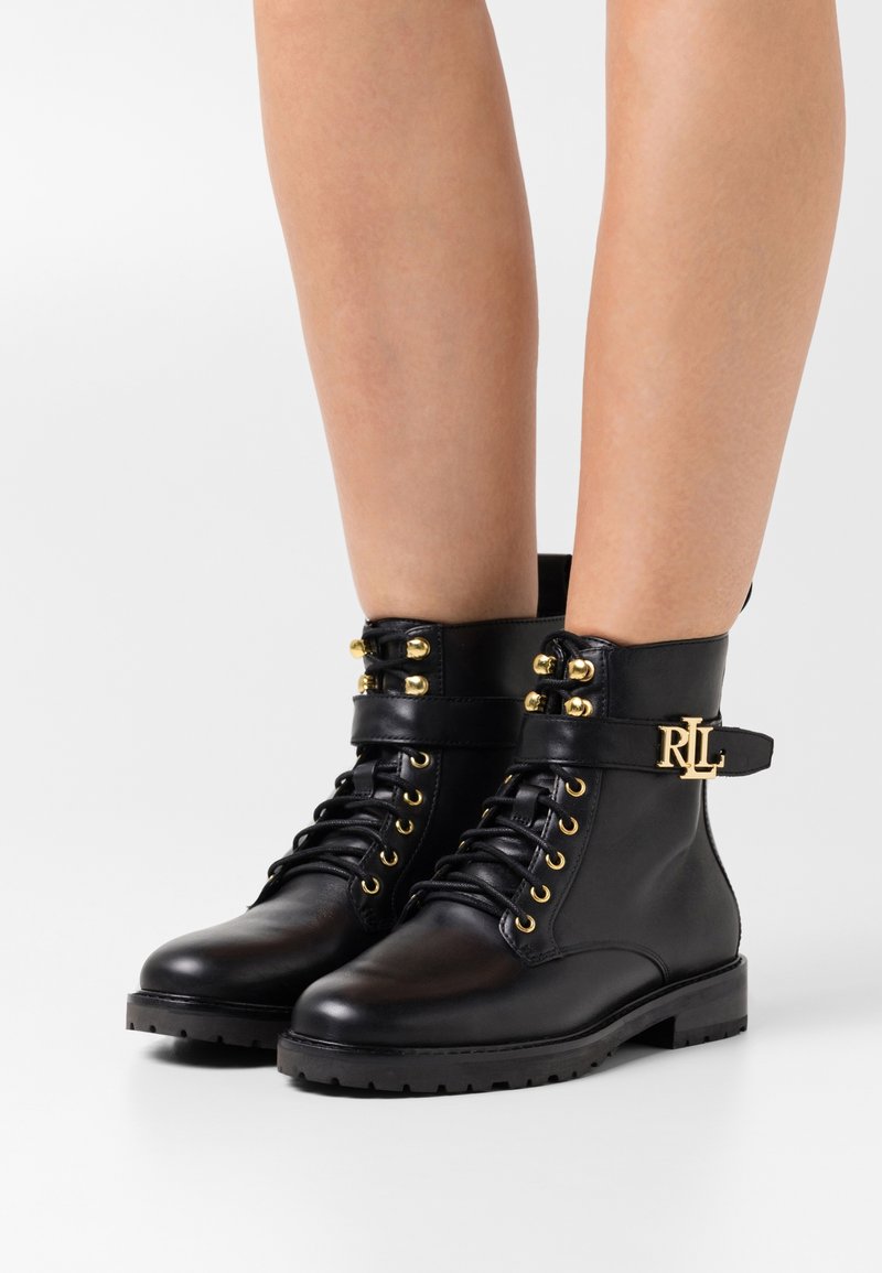 Botas de tobillo de cuero negro con herrajes dorados, punta redonda, diseño con cordones, y una correa con el logo "RL". Suela gruesa y texturizada.