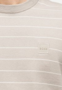 Μπεζ πλεκτό πουλόβερ με λεπτές λευκές οριζόντιες ρίγες και τετράγωνο μπαλώμα λογότυπο με την ένδειξη "BOSS" στο στήθος.