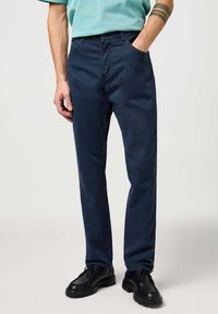 Pantalones azul marino fabricados en tejido duradero, con un diseño clásico de cinco bolsillos, corte recto y una textura sutil. Combinados con zapatos negros.