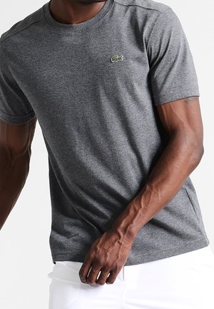 Mann trägt ein eng anliegendes graues Kurzarm-T-Shirt mit einem kleinen grünen Logo auf der Brust und weiße Hosen.