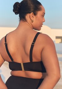 Top de bikini negru cu bretele reglabile și închizătoare aurie la spate. Textură netedă și design minimalist. Tonul pielii este vizibil.