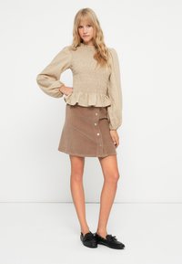 Blusa de color beige con fruncidos, de manga larga y dobladillo con volantes, combinada con una falda de pana marrón con botones en la parte frontal y un corte acampanado.