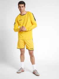 Gul atletisk skjorte med lange ærmer og rund halsudskæring, parret med matchende shorts. Har sorte detaljer og logo på brystet.