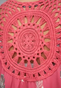 Koraalkleurige haakwerkdetails aan de achterkant met ronde motieven en open secties. De stof is lichtgewicht, met een zachte textuur en bloemrijke accenten aan de voorkant.