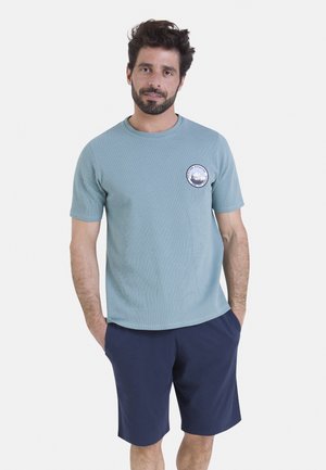 T-shirt bleu clair à manches courtes avec tissu texturé et logo circulaire sur la poitrine. Porté avec un short bleu marine foncé.