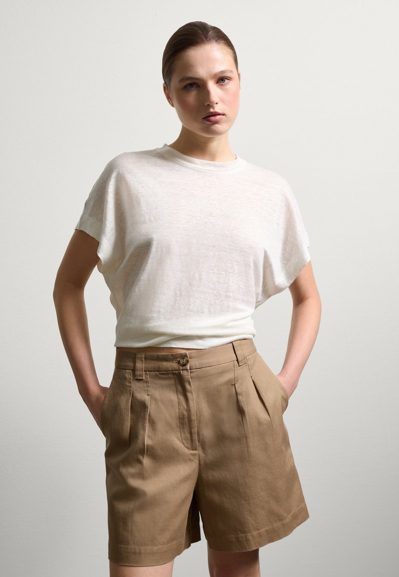 Helle beige Leinen-Shorts mit Falten und Seitentaschen, kombiniert mit einem lockeren, durchsichtigen weißen Baumwoll-T-Shirt. Minimalistisches Design, bequeme Passform.