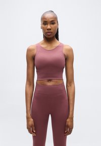 Αθλητική γυναίκα που στέκεται ακίνητη, φορώντας συνδυασμένο μοβ αμάνικο crop top και κολάν με ψηλή μέση, μπροστά σε ένα απλό φωτεινό φόντο.