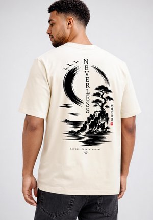 Man wearing a beige T-shirt with black art of a tree on a rock, birds, a crescent shape, and the word "NEVERLESS" in vertical text on the back.