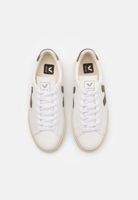 White leather sneakers with green suede accents, featuring white laces and a black and white logo on the tongue. Smooth texture and round shape.