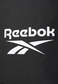 Zwart materiaal met een prominente witte Reebok-logo en een gestileerd wit swoosh-ontwerp. Het oppervlak heeft een textuur van stof.