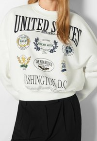 Biała bluza z grafiką, na której znajdują się nadruki, w tym "Stany Zjednoczone", różne emblemy oraz tekst "Waszyngton, D.C." Luźny krój z ściągaczami przy mankietach.
