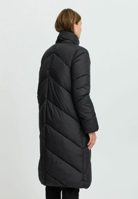 Abrigo acolchado negro con un cuello alto, con un patrón en forma de chevron, mangas largas y un diseño hasta el muslo, que destaca por su textura acolchada y suave.