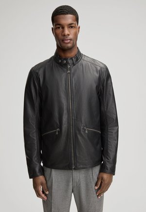 Man staat vooruit gericht en draagt een zwart leren jack met rits en opstaande kraag, gecombineerd met een grijze broek tegen een effen achtergrond.