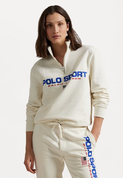 Hanorac crem cu fermoar, cu guler înalt, având inscripții albastre și roșii "POLO SPORT" și "RALPH LAUREN". Pereche de pantaloni de trening crem asortati.