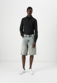 Camisa negra de manga larga combinada con pantalones cortos de mezclilla azul deslavado, con un ajuste relajado. Zapatillas blancas completan el atuendo casual.
