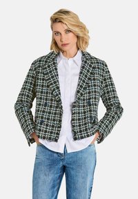 Blonde Frau, die ein schwarz-weiß kariertes Blazer über einem weißen Hemd und hellblauen Jeans trägt, die Hände in den Taschen, vor einem neutralen Hintergrund.