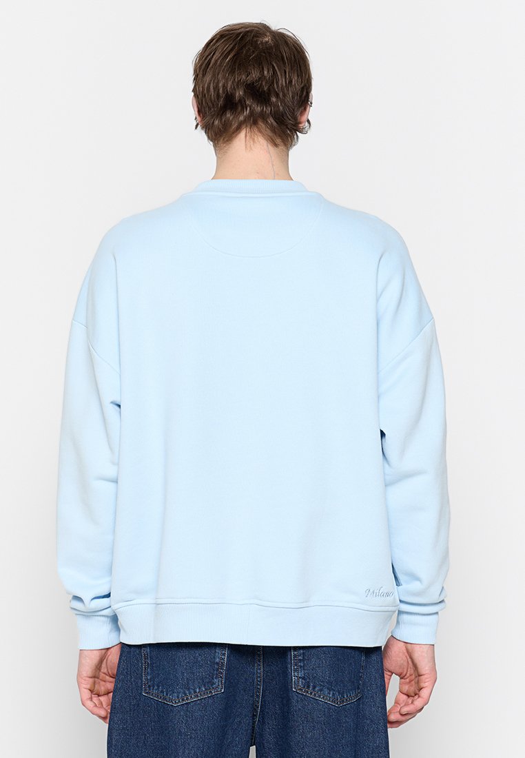 Persoon in een lichtblauw sweatshirt en donkerblauwe spijkerbroek, gezien van achteren tegen een effen lichte achtergrond.