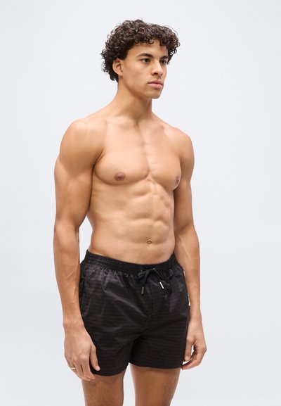 Αθλητικός άνδρας χωρίς μπλούζα με σγουρά μαλλιά, που φοράει μαύρα σορτς με κορδόνι, στέκεται μπροστά σε απλό ανοιχτό γκρι φόντο, στραμμένος ελαφρώς προς τα δεξιά.