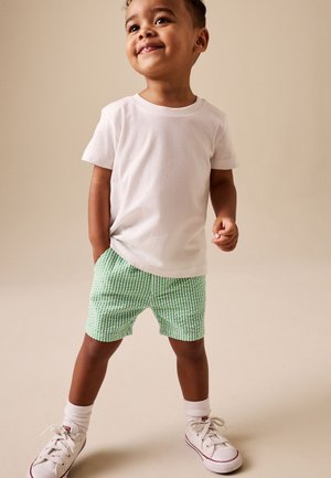 Lillebarn iført en hvid t-shirt, grønternet shorts, hvide sokker og hvide sneakers stående mod en beige baggrund.