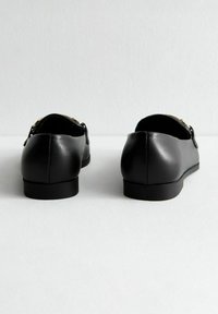 Zwarte leren loafers met een gladde textuur, lage hak en gouden hardware accenten aan de zijkanten. Weergegeven vanuit de achteraanzicht.