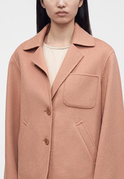 Femme portant un manteau en laine rose avec de grands revers, deux boutons marron, une poche poitrine et deux poches latérales sur un haut beige clair.