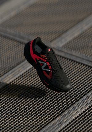 Sort og rød New Balance løbesko hængende over en metalrist med teksturerede mønstre.