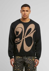 Schwarzer Pullover mit großem beigen Grafikanzeige auf der Vorderseite, ausgestattet mit gerippten Bündchen und Saum, kombiniert mit gemusterten Cargo-Hosen.