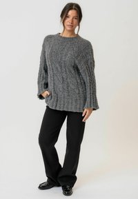 Maglione grigio chunky lavorato a maglia con modelli a treccia, maniche lunghe e orlo a coste, abbinato a pantaloni neri a gamba larga e scarpe lucide.