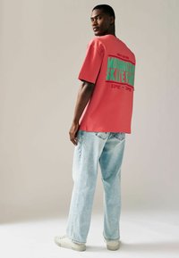 Rød oversized t-shirt med grøn grafisk tekst på ryggen, parret med lyseblå, løstsiddende jeans og cremefarvede sneakers.