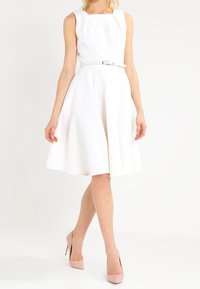 Robe blanche sans manches avec un décolleté carré et un tissu texturé. Présente une taille cintrée avec une fine ceinture et une jupe évasée.