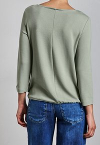 Ljusgrön långärmad topp med ett texturerat mönster och avslappnad passform, som är kombinerad med blå denimjeans med bakfickor.