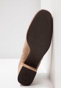 Bottine beige en daim reposant sur une surface blanche, semelle tournée vers l'avant, montrant une semelle noire lisse et un talon bloc avec logo.