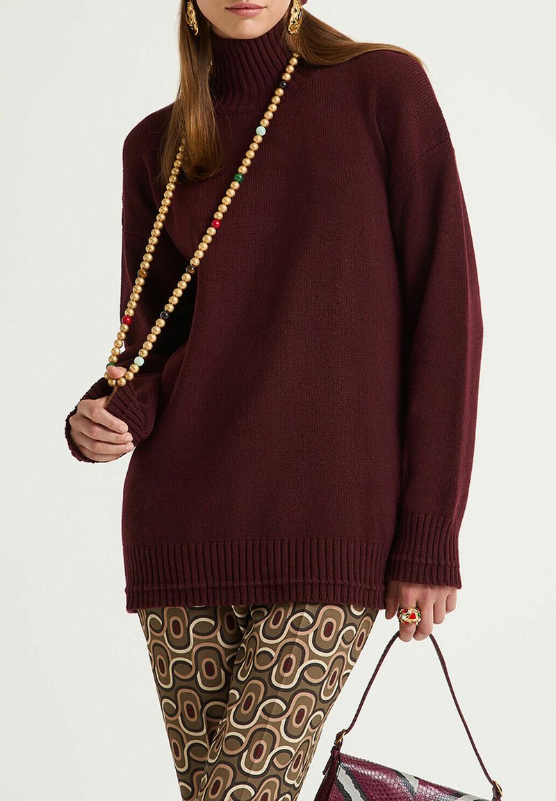 Maglione a dolcevita color bordeaux con orlo a coste; indossato con pantaloni marroni a fantasia e accessoriato con una collana di perline colorate e una borsa.
