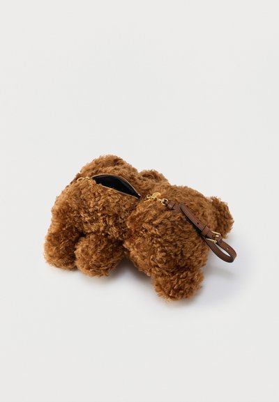 Καφέ τσάντα από ψεύτικη γούνα με μπούκλες, σε σχήμα σκύλου, με χρυστό φερμουάρ και καφέ δερμάτινο λουράκι καρπού, σε λευκό φόντο.