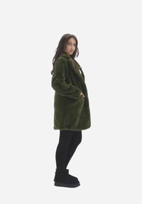 Cappotto in pelliccia sintetica verde con colletto alto, maniche lunghe e tasche laterali. Presenta una vestibilità ampia e una texture morbida e pelosa.
