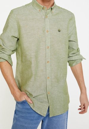 Homme portant une chemise boutonnée vert clair avec manches retroussées et un pantalon bleu, main dans la poche, debout devant un fond blanc.