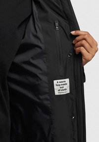 Schwarze Jacke mit einem glatten Innenfutter, ausgestattet mit einer Reißverschlusstasche und einem Etikett, auf dem steht: "Eine warme Umarmung aus Stoff."