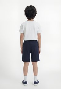 Παιδί που στέκεται πλάτη, φοράει λευκό κοντομάνικο πουκάμισο, ναυτικού χρώματος σορτς, λευκές κάλτσες και ναυτικού χρώματος αθλητικά παπούτσια σε απλό λευκό φόντο.