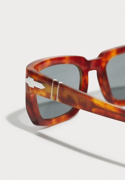 Óculos de sol tartaruga com armações grossas, lentes cinzentas e detalhe metálico nas hastes com um acento geométrico prateado.